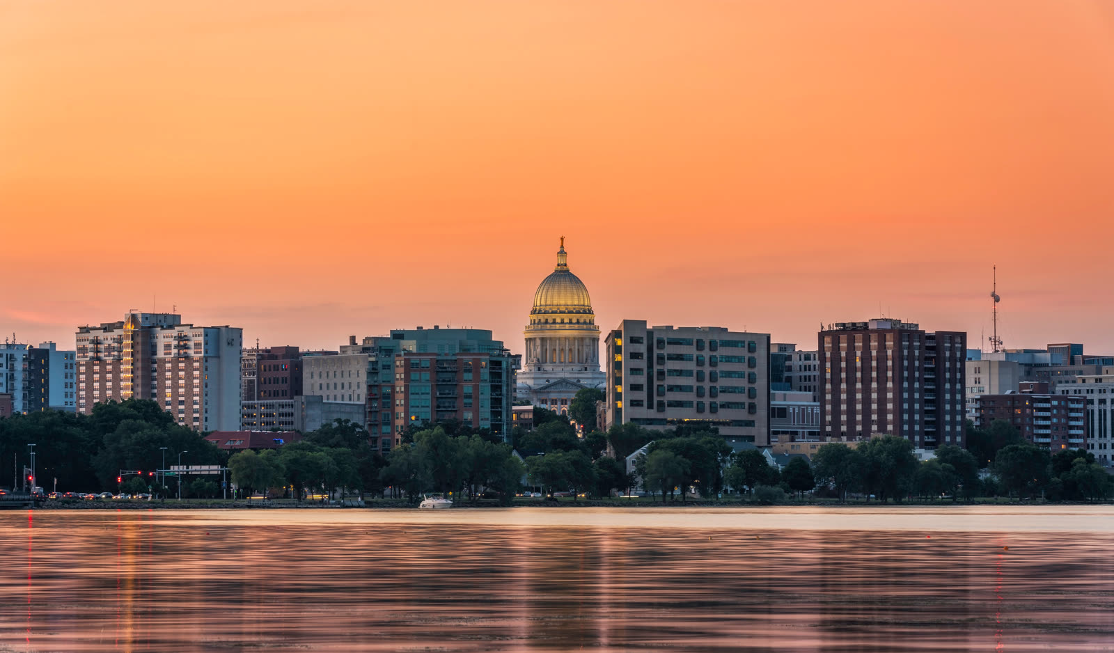 El edificio del Capitolio, con su cúpula, se eleva por encima de los rascacielos del centro de Madison, al otro lado de un lago tranquilo, bajo un cielo anaranjado al atardecer.