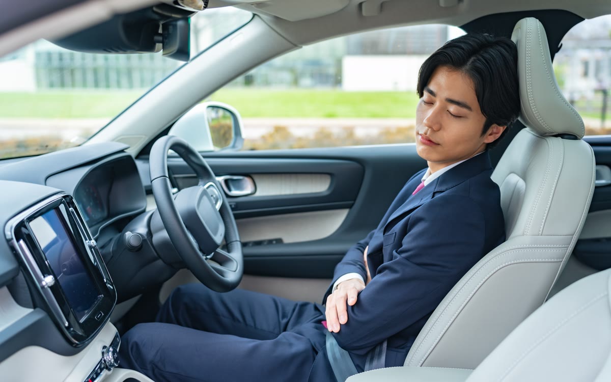 A man in a navy suit naps with his arms folded in the driver's seat of a car.
