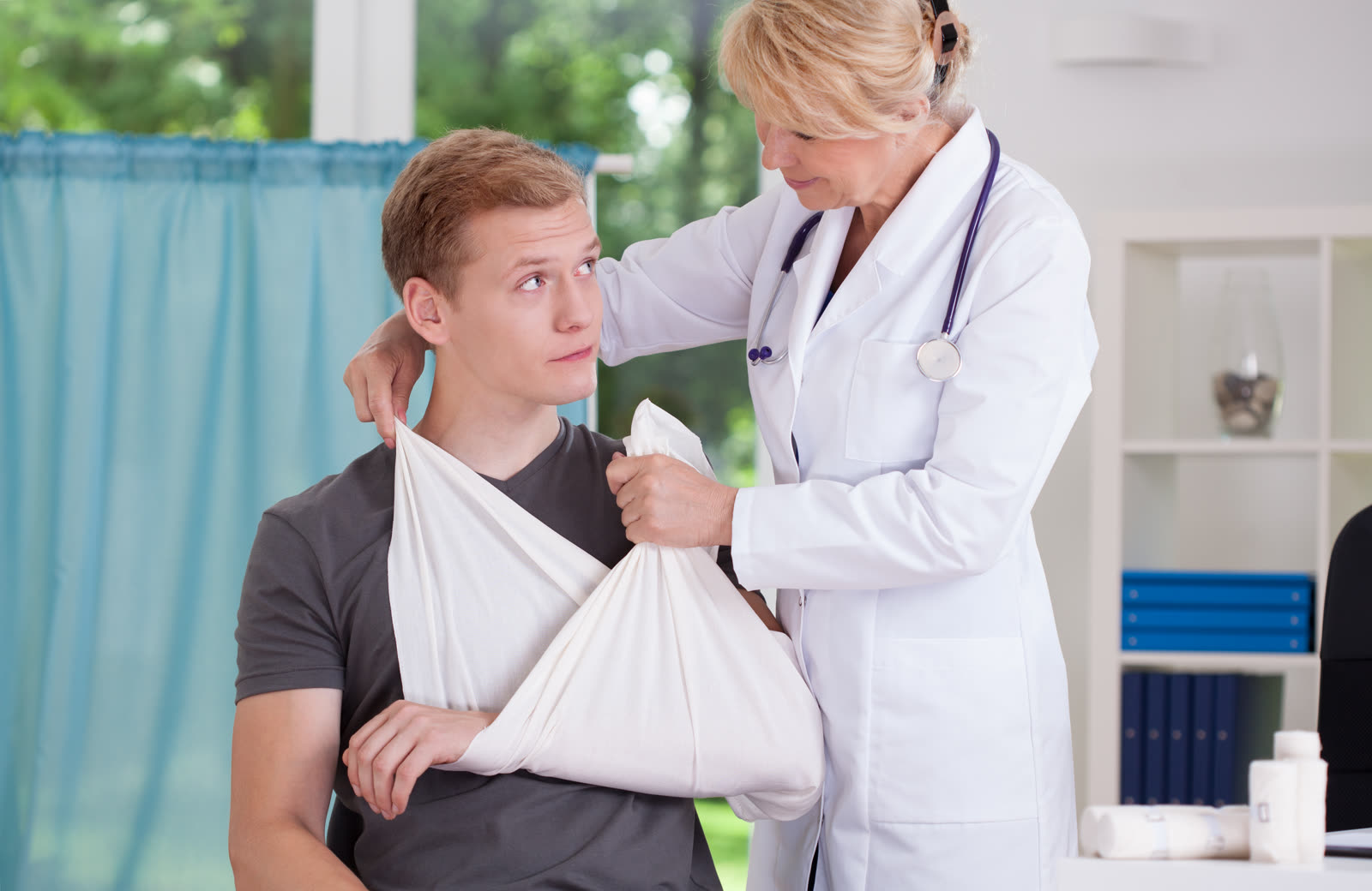 A doctor fits an arm sling on an injured young man at a clinic.