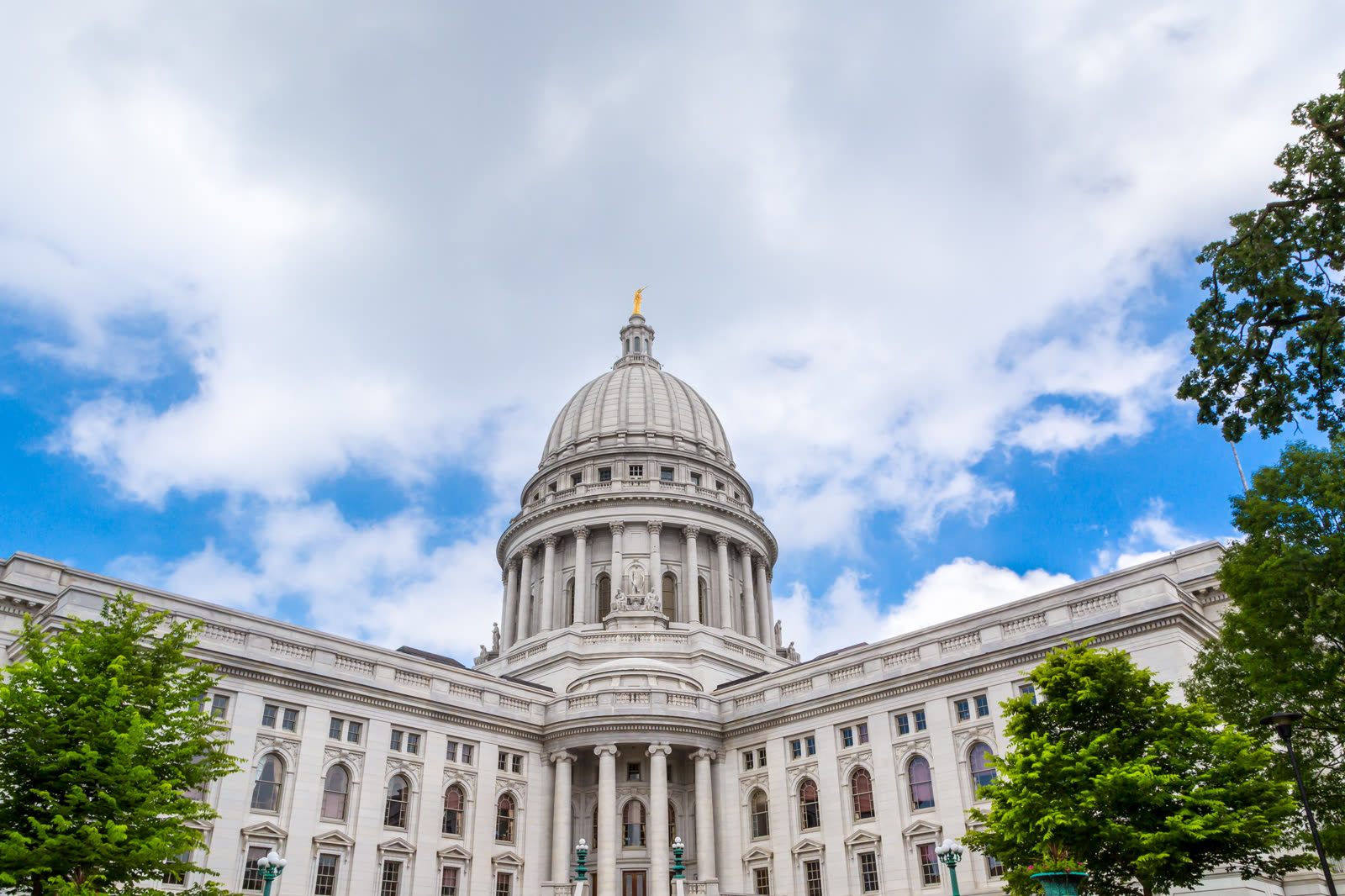 La cúpula de granito blanco del Capitolio del Estado de Wisconsin se eleva por encima de sus alas con columnas bajo un cielo azul salpicado de nubes, enmarcada por árboles verdes.